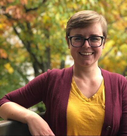 Katlyn Catron, a woman with pale pink skin and a short brown pixie cut, wearing black-rimmed glasses, a yellow shirt, and a maroon cardigan. She smiles and leans against a railing in front of a tree with vibrant fall foliage.